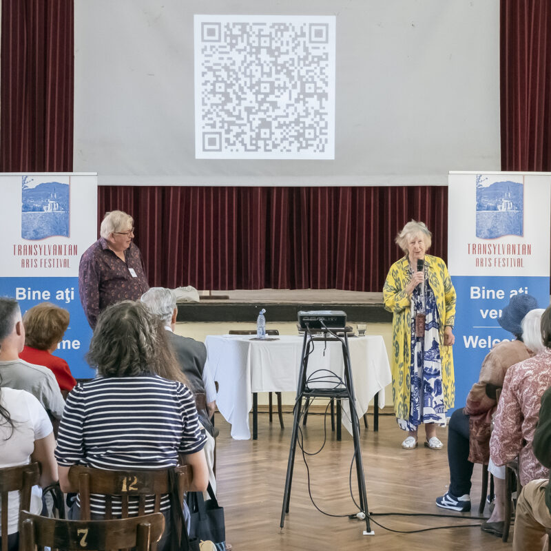 Festivalgründerin Lucy Abel Smith eröffnet das Transylvanian Arts Festival 2026 im Gemeindesaal von Reichesdorf. Links Professor Martyn Rady, der anschließend den ersten Vortrag hält.